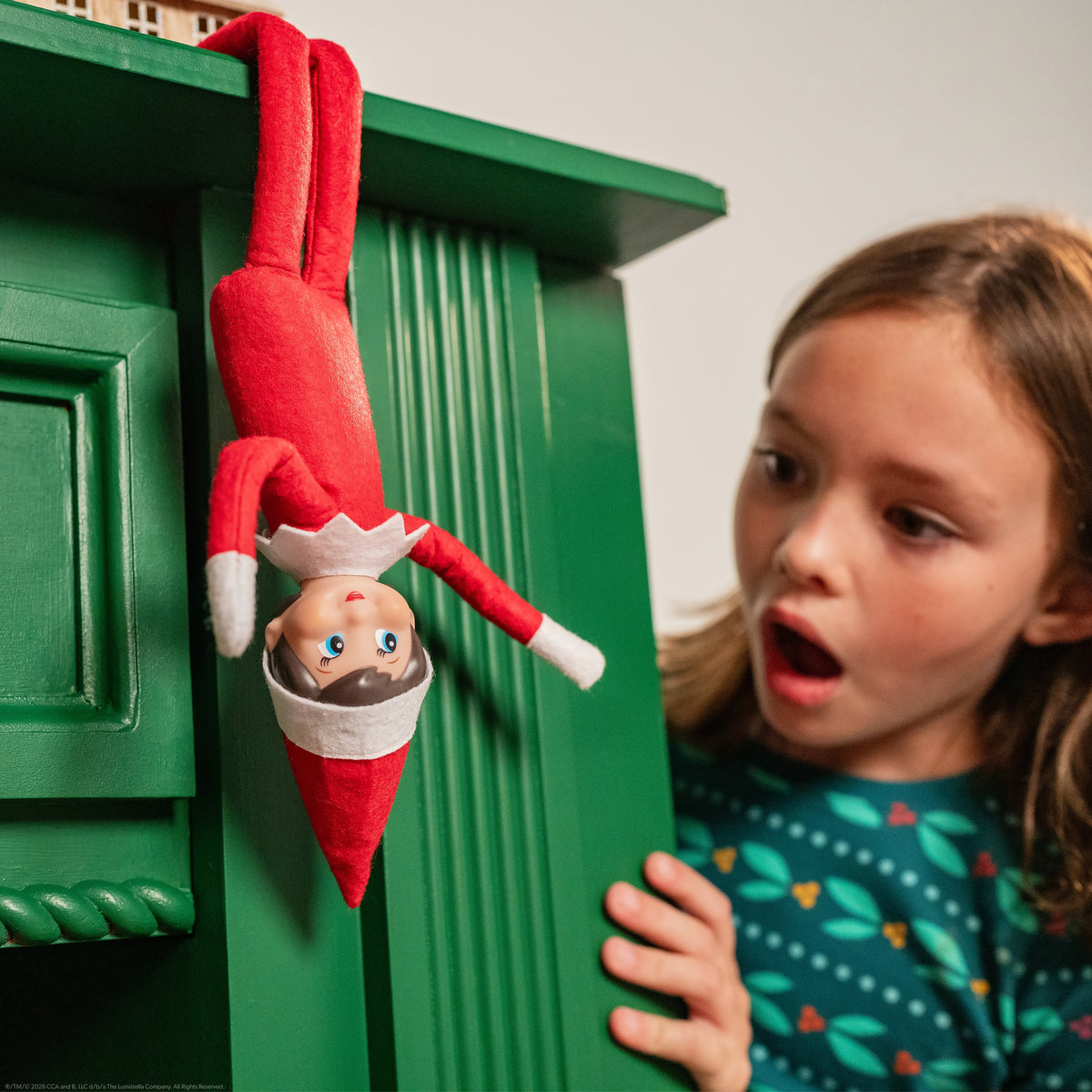 Poseable elf on the shelf in a red and white outfit hangs upside down from a green shelf, while a surprised child looks on.